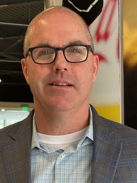 Professional headshot of Erich Manser. He is a middle-aged, white man with a shaved head and dark rimmed glasses. He is wearing a gray suit jacket, no tie, and a light-blue dress shirt.