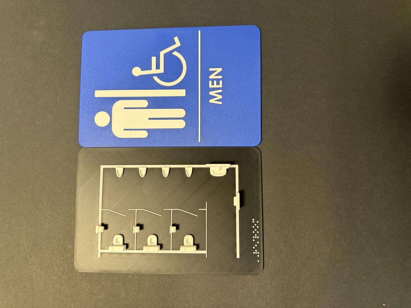 Blue ADA men's restroom sign mounted above a 3D-printed tactile map of the restroom layout.