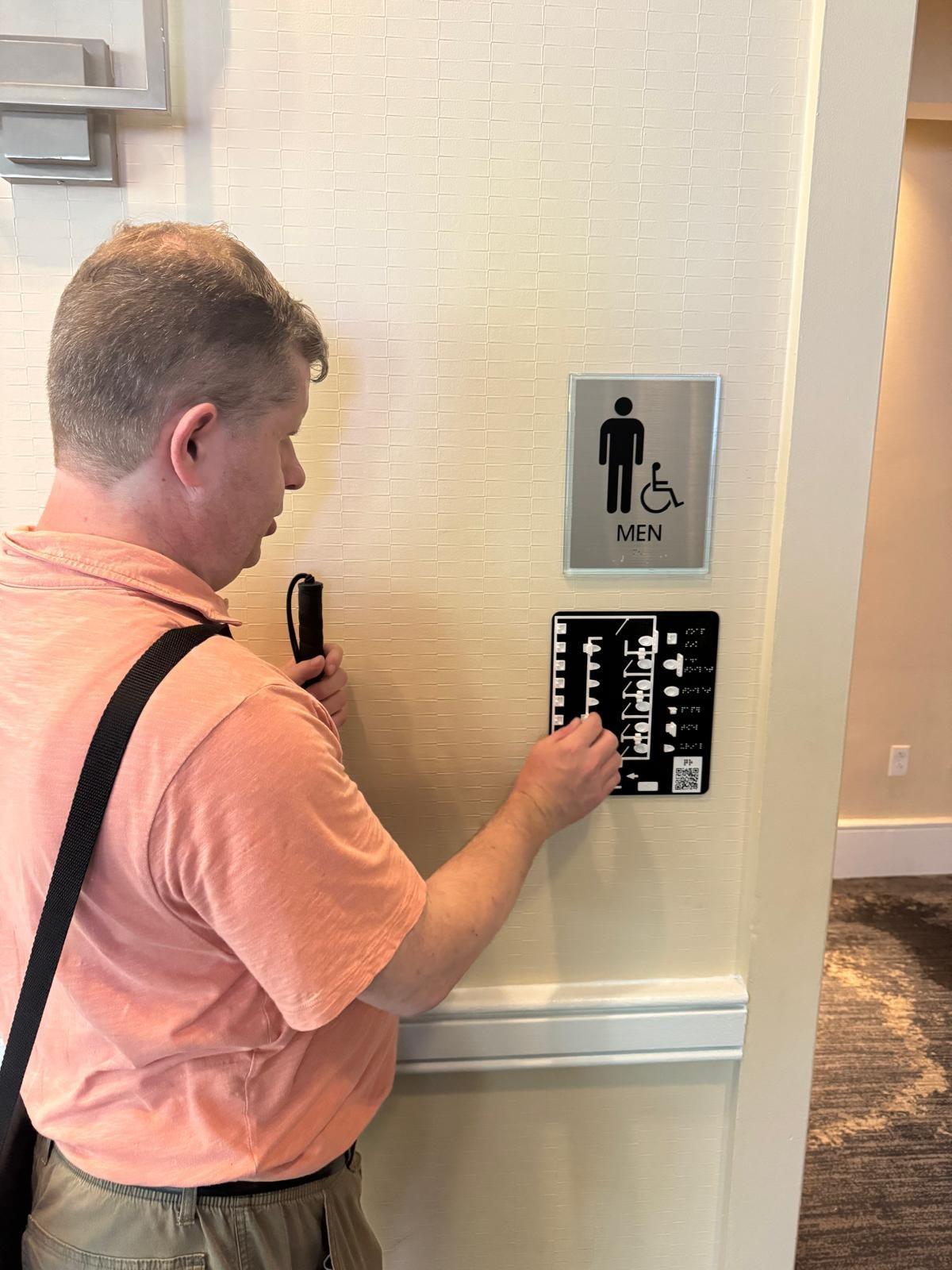 Tom Babinszki, holding a white cane, reading a tactile restroom map by touch. The map is mounted on a wall below a men's restroom sign.